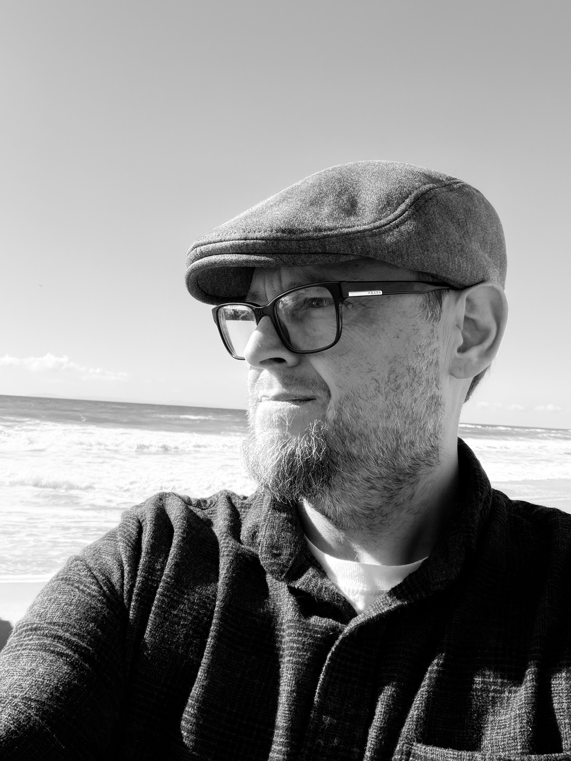 Black-and-white coastal portrait of Jason Alves in a flat cap and glasses